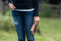 Hand in jeans pocket, holding a bible outside.