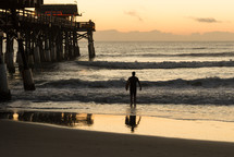 Silhouette of a man carrying a surfboard walking out in to the ocean waves at daybreak near a pier.