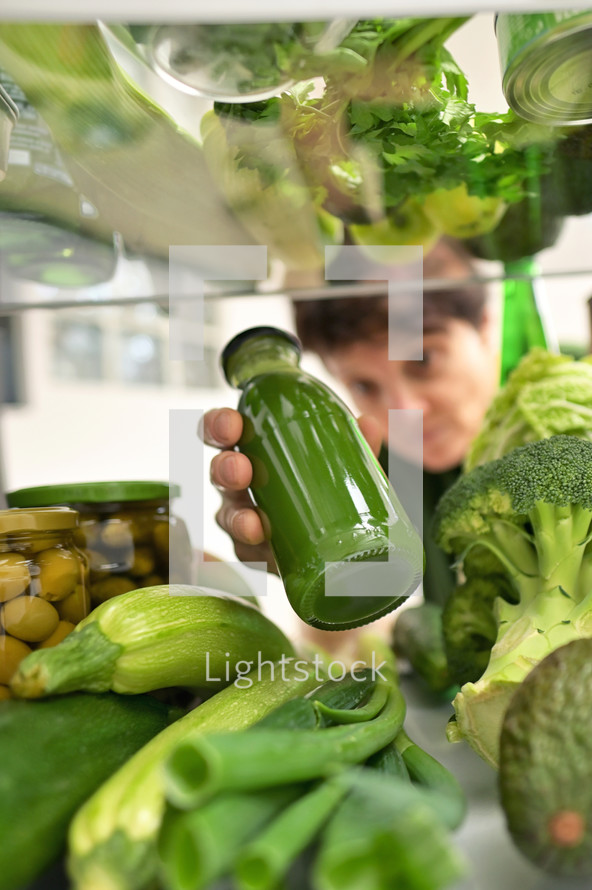 Woman choosing a bottle with green juice from refrigerator, View through fridge