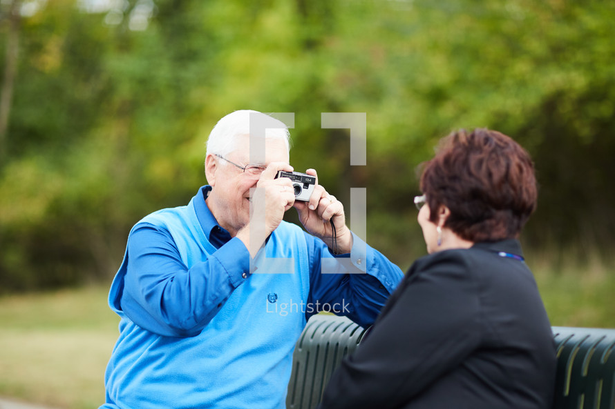 elderly couple sitting on a park bench taking pictures with a camera 