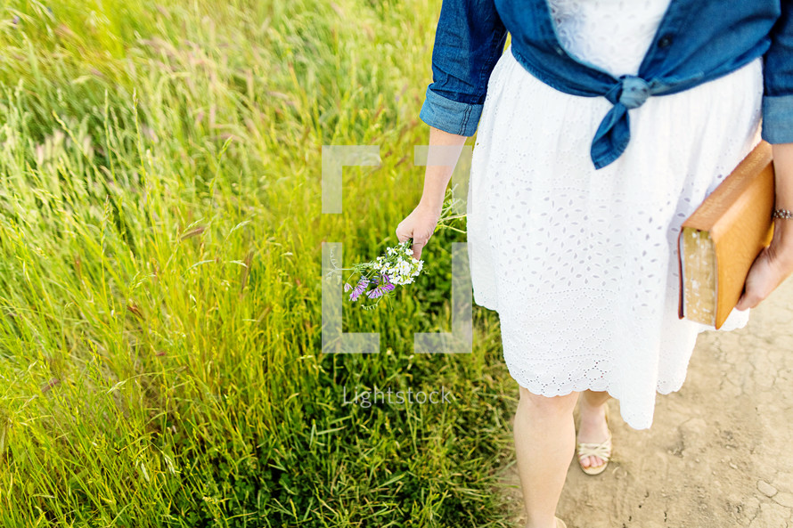 Woman walking in field holding a bouquet of flowers and a Bible.