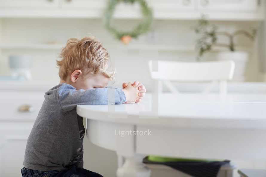 toddler boy praying at a kitchen table 