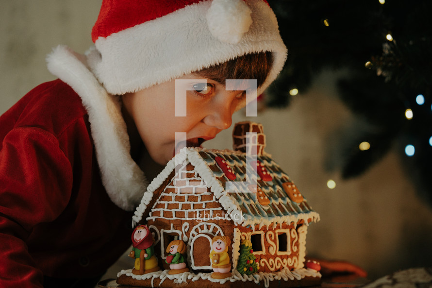 Cute little boy in Santa Claus costume eating gingerbread house on Christmas tree background. Home celebration, decoration with lights. Happy childhood, kids, lovely son. High quality photo