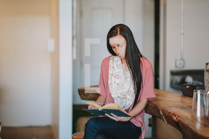 a young woman sitting on a stool in a kitchen reading a Bible 