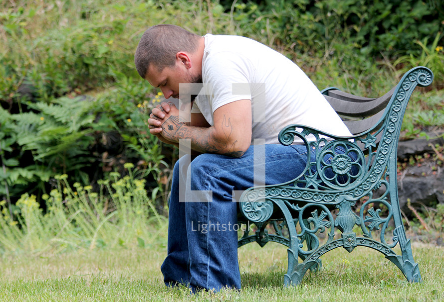 man with tattooed hands praying sitting on a park bench