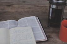 An open Bible and notebook next to an orange coffee mug and french press