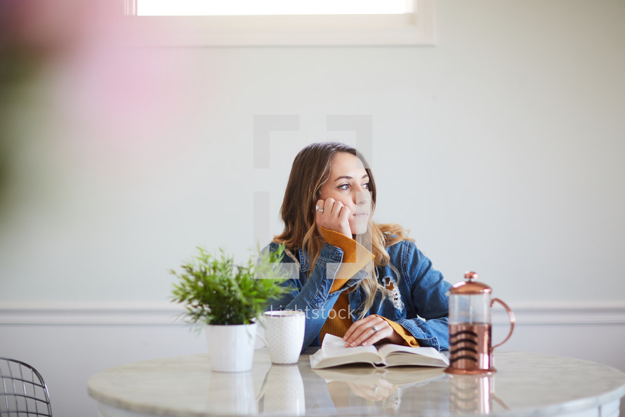 a woman reading a Bible sitting at a kitchen table and staring off in thought 