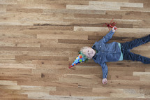 a toddler boy lying on a wood floor wearing a party hat. 