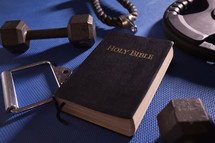 Holy Bible surrounded by dumbbells weights and other exercise equipment