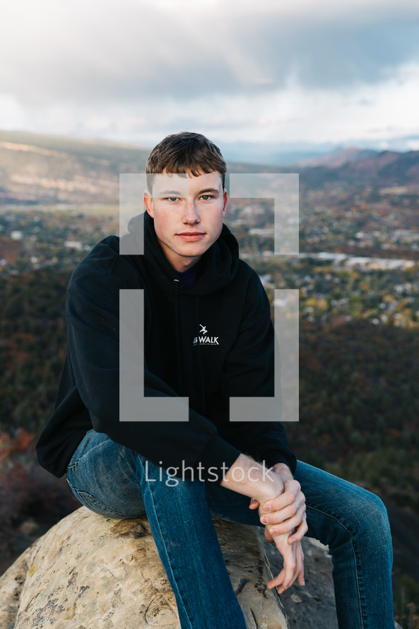 portrait of a young man in a hoodie sitting on a rock with valley in background 