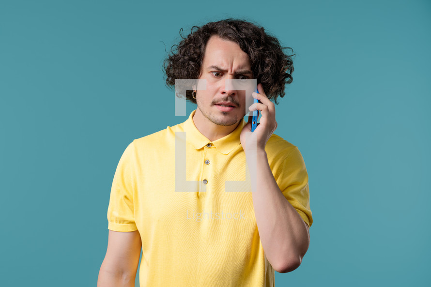 Annoyed young man quarreling while talking on mobile phone. Angry dude, stress. Blue studio background. Conflict, dispute, negative. High quality 