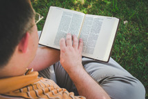 a man sitting in the grass on a sunny day reading a Bible God's Word with a view of him
from above. 