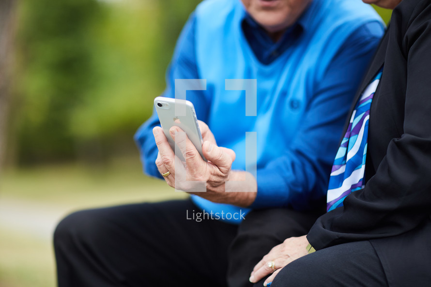 elderly couple sitting on a park bench looking at a cellphone screen 