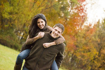 Man carrying a woman on his back through a field with fall foliage.