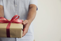 A man holding out a gift wrapped in brown paper and a red bow.
