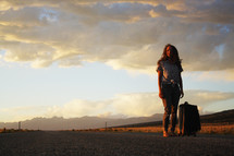 a young woman standing in the middle of a road with a suitcase at sunset 