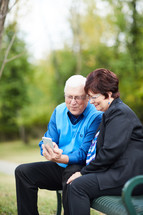 elderly couple sitting on a park bench looking at a cellphone 