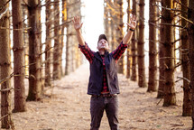a man standing outdoors in the woods with hands raised 