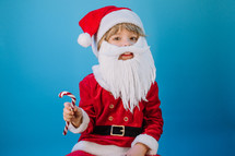 Funny boy in Santa Claus hat and sweet caramel cane on blue studio background. Christmas celebration. Happy childhood, kid, lovely son. High quality photo