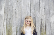 A little girl with glasses and blonde hair stands against a wooden fence.