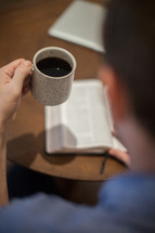 A young man sitting at a table reading the Bible holding a cup of coffee