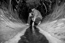 Man kneeling and touching the water flowing through a sewer drain pipe painted with graffiti.