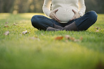 torso of a pregnant woman sitting in the grass with her hands on her belly