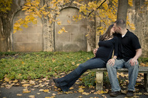 pregnant woman kissing a man sitting on a park bench