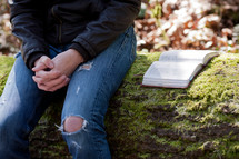 Man in ragged jeans sitting on a mossy log, hands folded, with open Bible.