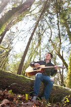 man sitting on a fallen tree playing a guitar in a forest