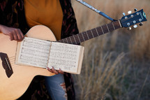 a woman standing in a field with a guitar and sheet music 