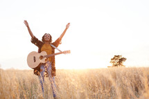 a woman standing in a field with a guitar with hands raised 