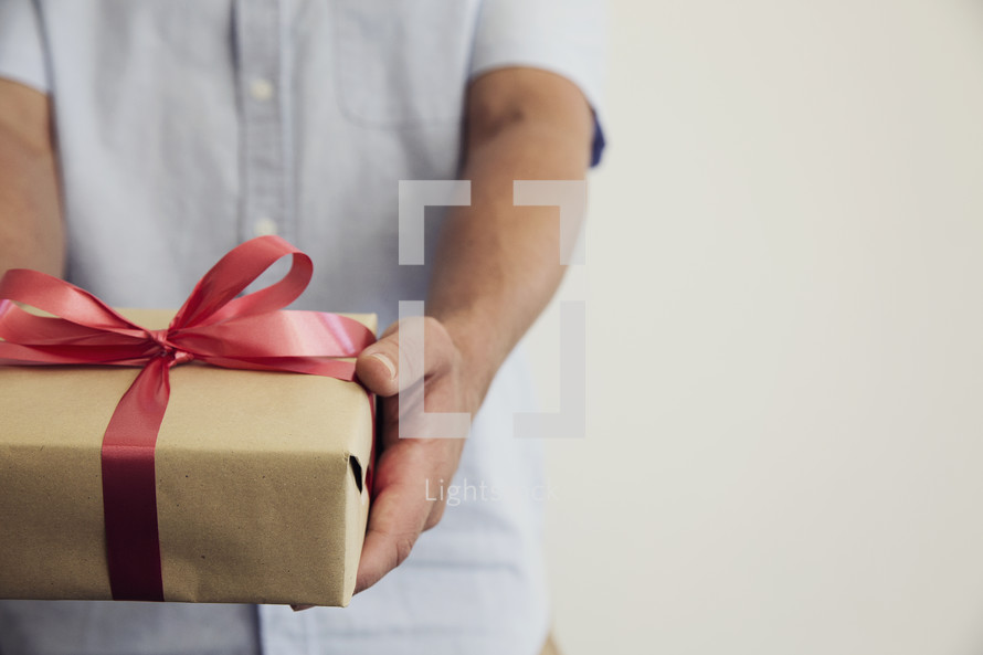 A man holding out a gift wrapped in brown paper and a red bow.