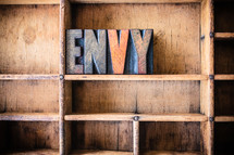 Wooden letters spelling "envy" on a wooden bookshelf.