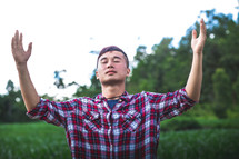 young man in a plaid shirt standing outdoors with hands raised 