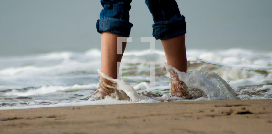 Child standing in the sand on a beach with ocean water rushing over feet.