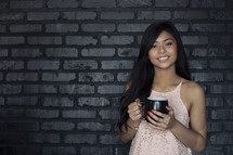 A smiling young woman holding coffee cup and standing against a gray brick wall.