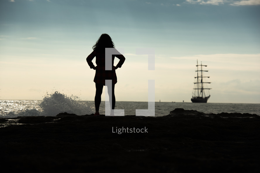silhouette of a woman standing on an ocean shore looking out at a ship 