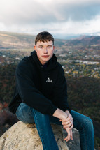 portrait of a young man in a hoodie sitting on a rock with valley in background 