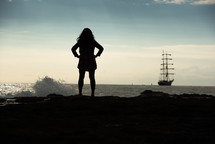 silhouette of a woman standing on an ocean shore looking out at a ship 