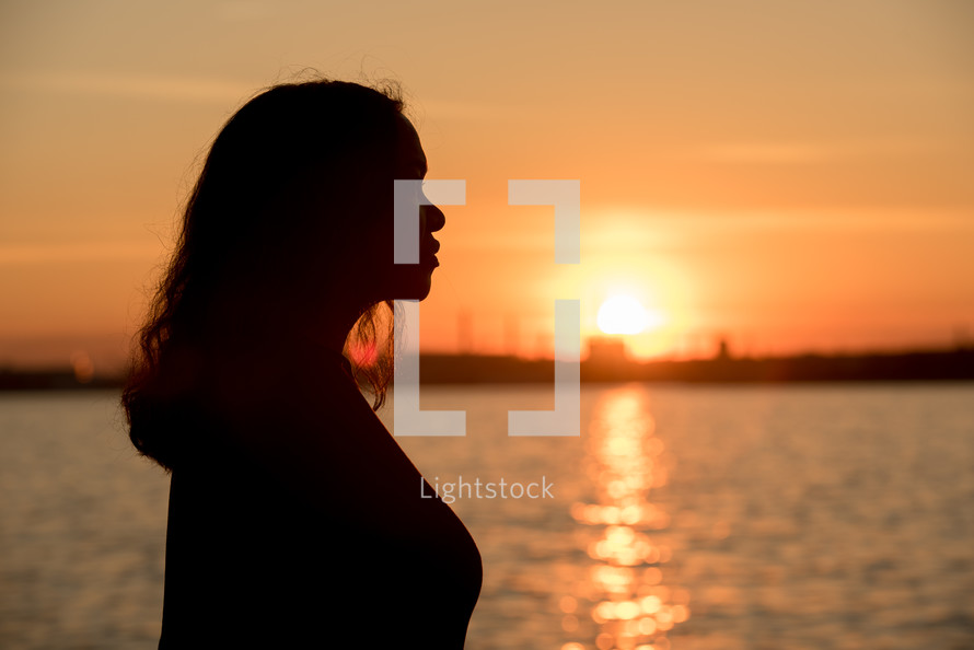 silhouette of a woman standing by water at sunset 