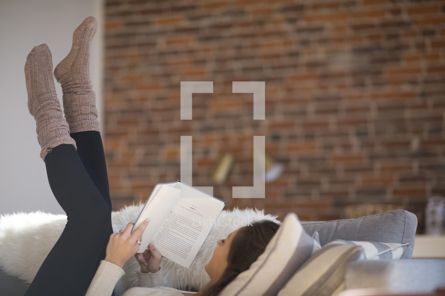 a woman lying on a couch with her feet in the air reading a book