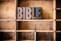 Wooden letter spelling "Bible" on a wooden bookshelf.
