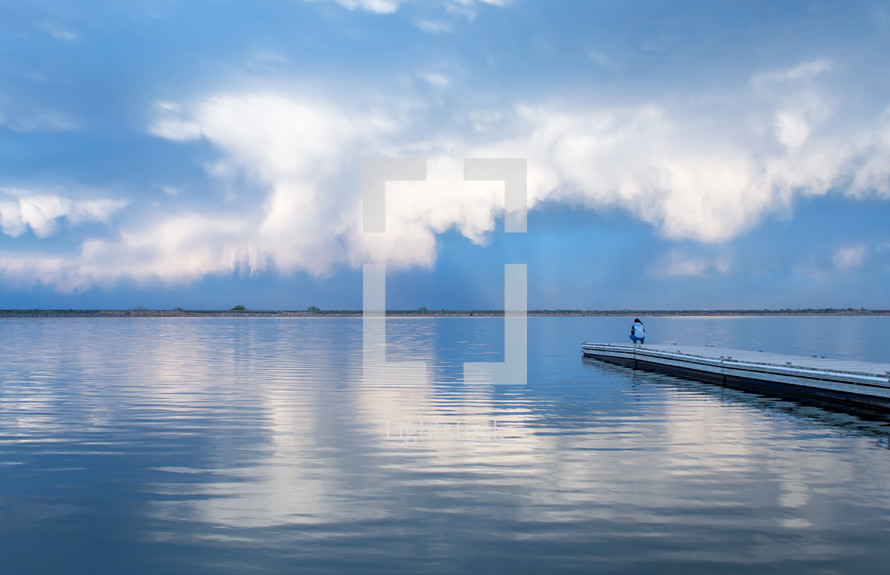 man standing at the end of a dock on a lake under a cloudy sky