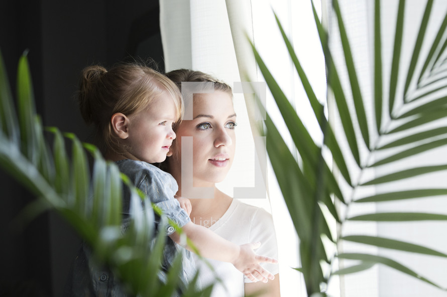 a mother and toddler daughter standing at a window 