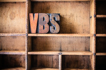 Wooden letters spelling "VBS" on a wooden bookshelf.