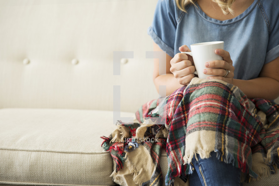 a woman sitting on a couch with a plaid blanket in her lap holding a mug 
