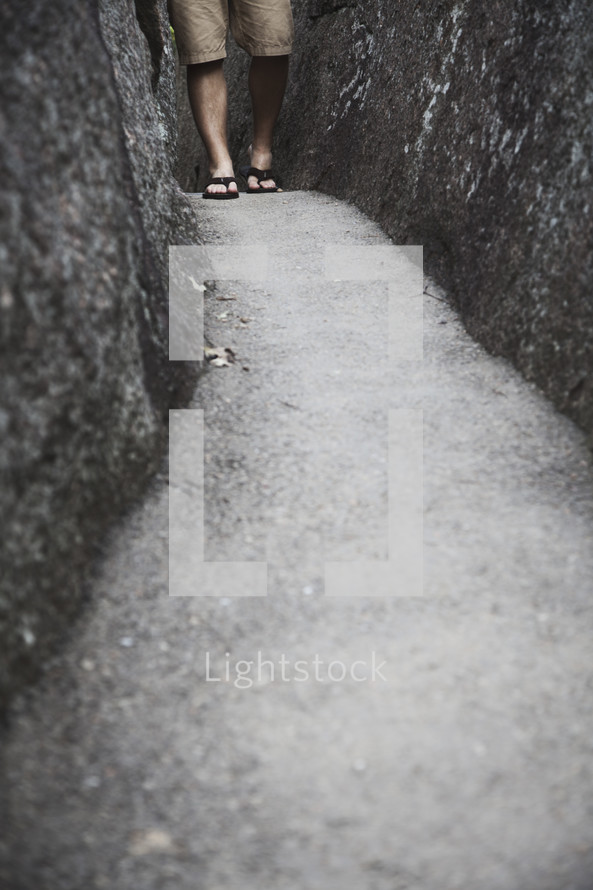 Feet in sandals walking on a paved path between two boulders.