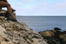 a man in boots sitting on rocks along a shore 
