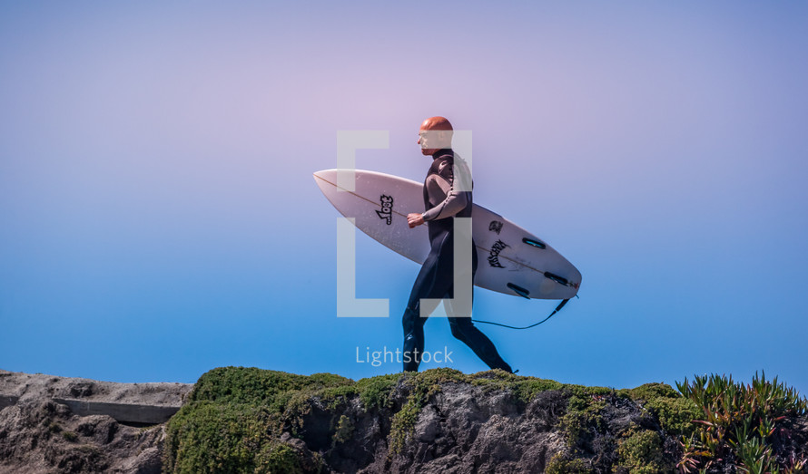 a surfer carrying his surfboard on a rocky shore 